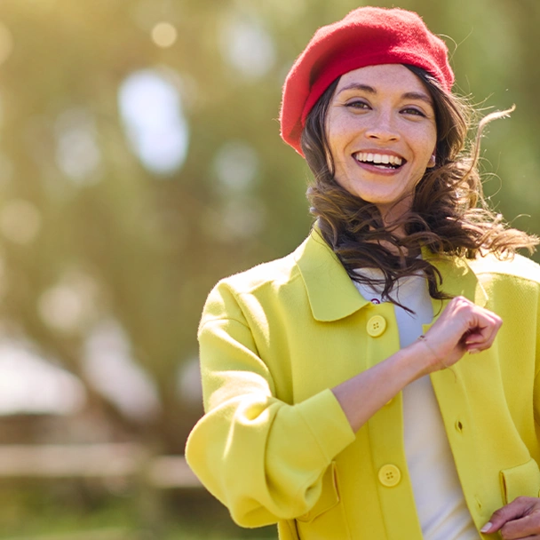 Une femme en veste jaune et béret rouge sourit en marchant dans un parc