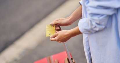Une personne tient en mains une Mastercard gold by Cofidis à côté d'une valise