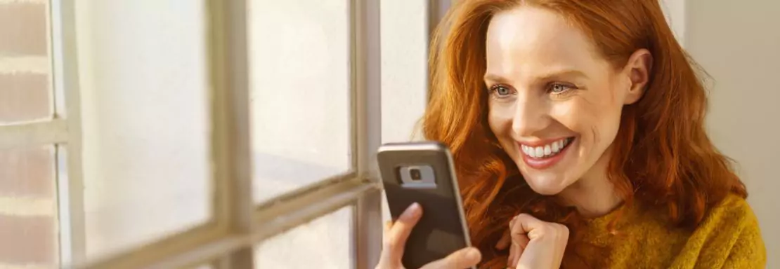 Vrouw glimlacht naar smartphone bij raam, zonnige dag